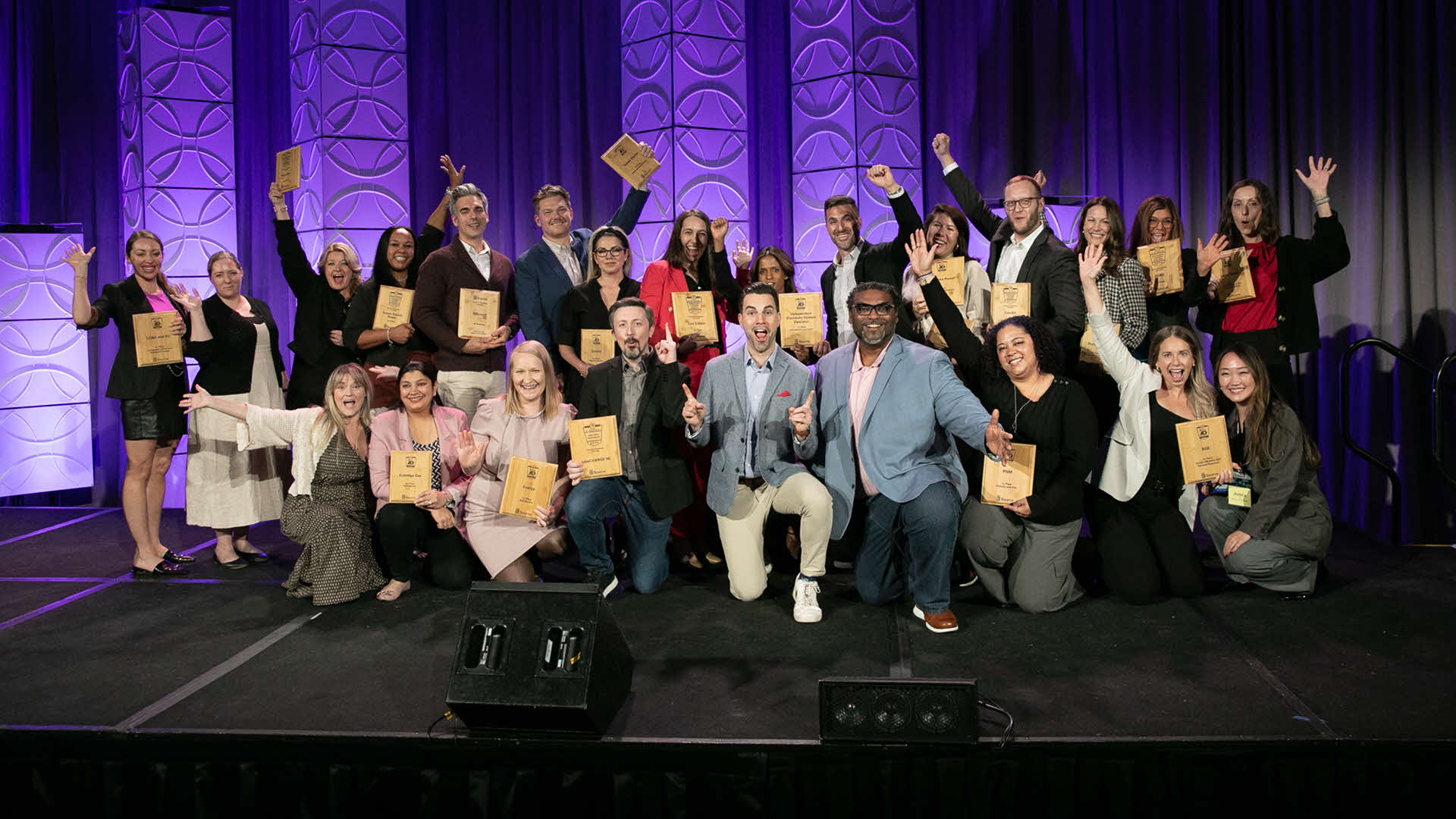Group of people on stage holding award plaques in front of a purple backdrop