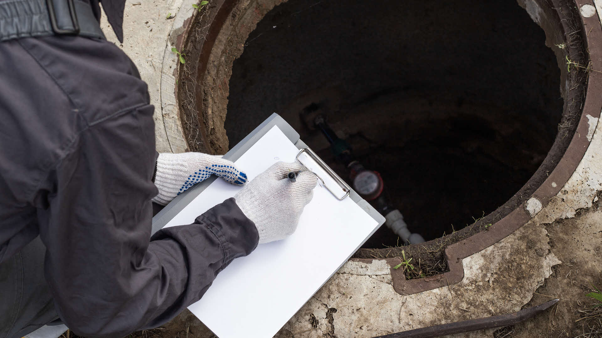Person writing on a clipboard while inspecting an open manhole