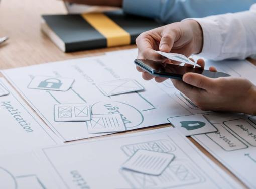 Person holding a smartphone and paper cutouts over application design diagrams on a desk.