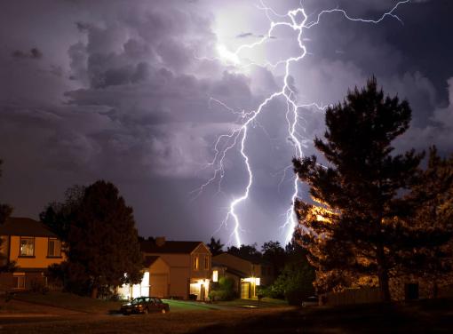Bright lightning bolts strike behind a suburban neighborhood at night, illuminating dark storm clouds and silhouetted trees