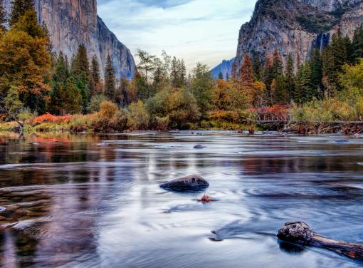 A calm river flows through a valley surrounded by autumn-colored trees, with tall granite cliffs rising in the background.