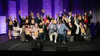 Group of people on stage holding award plaques in front of a purple backdrop